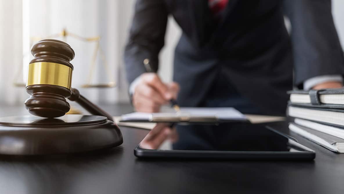 close up of a desk with a gavel and tablet in the foreground, a judge signing a paper in the background